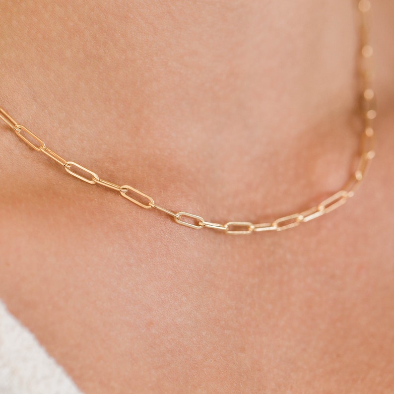 May include: A delicate gold-colored chain necklace with a paperclip link design. The necklace is shown on a person's neck, highlighting the chain's subtle elegance. The links are rectangular and evenly spaced.