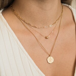 May include: A layered gold necklace set. The top layer features a paperclip chain, followed by a delicate chain with a small bead, and a pendant necklace with a detailed coin design. The necklaces are worn on a person.