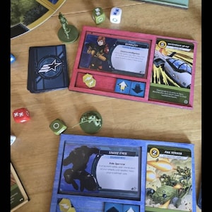 May include: A blue and red wooden game board with two card slots and two dice slots. The cards are from the G.I. Joe game and feature characters Scarlett and Snake Eyes. The text on the cards reads "Scarlett, Commander in Chief" and "Snake Eyes, Classified".