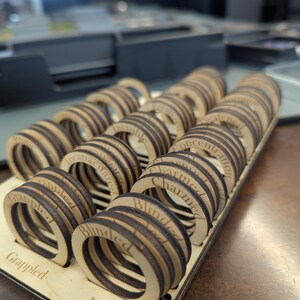 May include: A wooden tray holds multiple laser-cut wooden rings with various engraved words, including "Blinded," "Charmed," and "Grappled." The rings have a layered design with dark brown and light brown wood tones. The tray is also made of light-colored wood.
