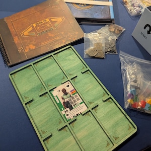 May include: A board game setup featuring a green wooden organizer with card slots, a card with a character illustration, and various game components. There are two books, one with the text "BOOK OF VANTAGES", and several small bags of game pieces.