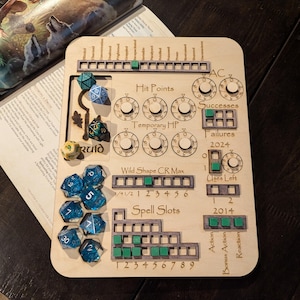 May include: A wooden tabletop gaming tracker with a green and gray color scheme. It features a variety of dials, squares, and spaces for tracking character stats, including hit points, temporary hit points, successes, failures, spell slots, and wild shape CR max. The tracker also includes spaces for dice, with several blue dice visible in the image.