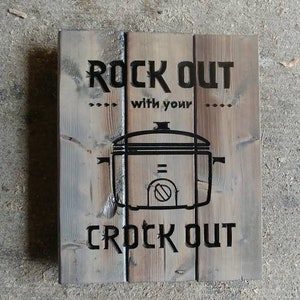 Peut inclure: Une pancarte en bois avec le texte "Rock Out with your Crock Out" en lettres noires. Une silhouette noire d'une mijoteuse est au centre de la pancarte.