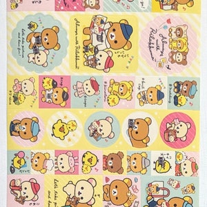 May include: A sheet of colourful stickers featuring the Rilakkuma bear character in various poses. The stickers have a yellow, pink, and blue colour scheme, with text that says "Always with Rilakkuma" and "Let's take pictures and have fun!"