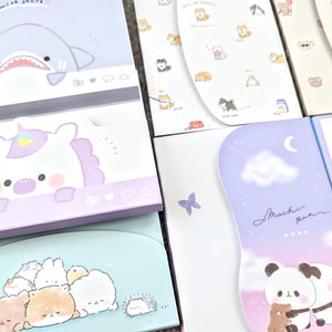Könnte beinhalten: Ein Stapel bunter Notizblöcke mit niedlichen Tierillustrationen. Der oberste Block zeigt einen blauen Hai mit dem Text "wish you animal". Der zweite Block zeigt ein lila Einhorn mit dem Text "love". Der dritte Block zeigt eine Gruppe von braunen und weißen Tieren. Der vierte Block zeigt einen Pandabären mit dem Text "Muchie pun". Der fünfte Block zeigt eine Vielzahl von bunten Aufklebern mit Tieren und Text.