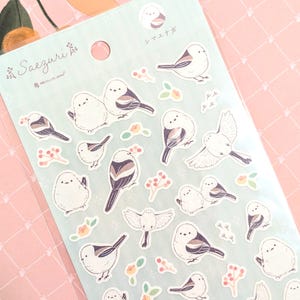 Może przedstawiać: Arkusz naklejek z uroczymi ilustracjami sikorek, małych ptaków. Naklejki są na jasnoniebieskim tle z białymi i żółtymi akcentami kwiatowymi. Na arkuszu nadrukowano tekst "Saezuri" i "シジュウカラ".