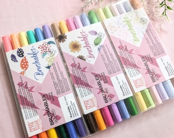 May include: Three sets of ZIG Brushables markers in clear plastic packaging. Each set contains a variety of colors, including pastels and vivid hues. The packaging features floral designs and the text "Brushables" and "Kuretake Est. 1902".