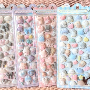 May include: Four sticker sheets with the text "FUWAFUWA" and "FUWAFUWA DROP". Each sheet features various pastel-colored, puffy stickers. Designs include bows, bears, cats, penguins, and other cute shapes. The stickers are arranged on clear plastic backing.