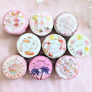 Puede incluir: Colección de nueve rollos de cinta washi decorativa. Cada rollo presenta un diseño único, incluyendo patrones florales, ilustraciones de ciervos y gráficos de verano. Los rollos son principalmente rosas y blancos con detalles dorados, perfectos para manualidades y scrapbooking.