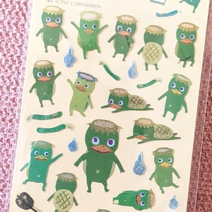 May include: Sheet of stickers featuring cartoon-like green creatures with glittery accents. The stickers depict various poses, including standing, waving, and lying down. Additional stickers include blue teardrop shapes and green curved lines.