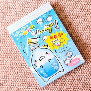 May include: A notepad with a light blue cover featuring a cartoon illustration of a soda bottle with a face. The notepad has Japanese text and a 500ml measurement. The background is a pink, textured surface.