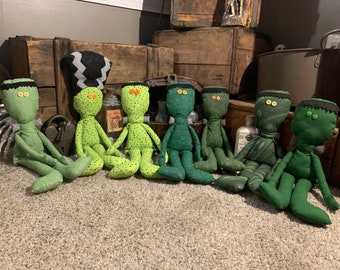 May include: A collection of seven fabric dolls in various shades of green, each featuring button eyes and stitched hair details. The dolls are crafted from different green textiles, including solid, dotted, and patterned fabrics, and are seated in a row.