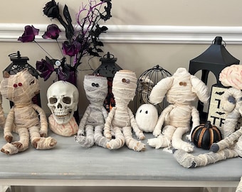 May include: A collection of Halloween-themed decor including several fabric mummy dolls wrapped in beige gauze, a human skull, a white ghost figure, a black witch hat, a black lantern, a small pumpkin, and a brain-shaped prop.