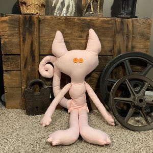 May include: A light pink cat-shaped plush doll with button eyes and a curled tail. The doll has long arms and legs, and is wearing a small pink garment. The doll is sitting in front of a wooden crate.