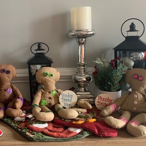 May include: Three gingerbread people dolls with different colored buttons and embroidered details. The dolls are sitting on a red and green patterned surface with a wooden sign that says "Wobble Bread Man" and another sign that says "Monster Bread".