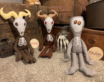 May include: Three plush dolls featuring skeletal designs. Two dolls have horned skulls with white faces and ribcage details. The third doll is gray with button eyes and a ribcage patch. Two circular wooden signs are labeled Wendigo and Windigo.