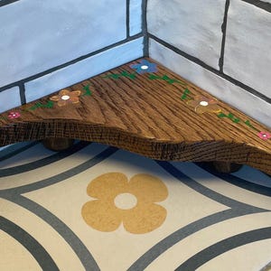 May include: A wooden corner shelf with a natural finish, adorned with hand-painted flowers in pink, white, and blue. The shelf is supported by three small, rounded feet and is designed to fit into a corner space.