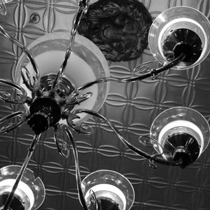 May include: A silver chandelier with multiple light bulbs hangs from a ceiling with a decorative pattern. The chandelier has a central hub with multiple arms extending outwards, each ending in a light bulb fixture.