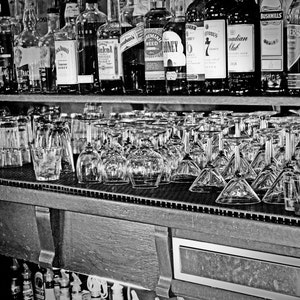 May include: A shelf with a variety of liquor bottles and glasses. The bottles are arranged in two rows, with the top row containing clear glass bottles with labels and the bottom row containing clear glass bottles with labels. The bottles are all different sizes and shapes. The glasses are arranged in a row at the bottom of the shelf. The glasses are all clear and have different shapes. The shelf is made of wood and has a dark brown finish.