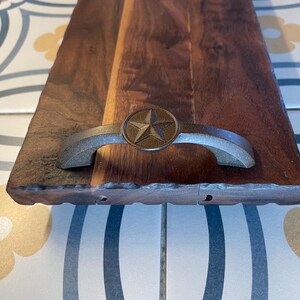 May include: A wooden serving tray with a live edge and a metal handle with a star design. The tray is made of walnut wood and has a natural finish.