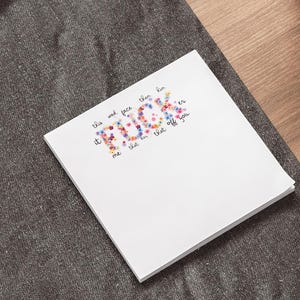 Peut inclure: Un bloc-notes blanc avec le mot "FUCK" écrit en lettres fleuries multicolores. Le bloc-notes est posé sur une surface gris foncé, avec un ordinateur portable et un stylo en arrière-plan. Une surface en bois et une plante en pot sont également visibles.