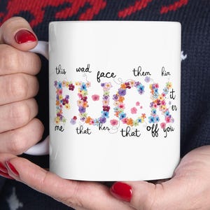 May include: White ceramic mug with the word "FUCK" spelled out in colorful floral letters. Surrounding text includes phrases like "this wad face them him it", "me that her that off you". The mug is held by a person.