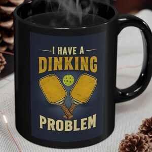May include: A black ceramic mug featuring a graphic with two yellow pickleball paddles and a yellow ball. The text reads I HAVE A DINKING PROBLEM in yellow capital letters. The mug has a handle on the right side and a dark blue rectangular background.
