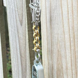 May include: Close-up of a silver and gold-toned metal chain with a clasp and decorative elements. The chain is hanging in front of a wooden fence, with the metal reflecting the sunlight.