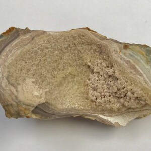 Puede incluir: Primer plano de una geoda de ágata de color marrón claro, beige y blanco, con una forma irregular y rugosa. La geoda tiene un patrón de capas y bandas y un pequeño grupo de cristales blancos.