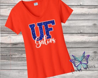 May include: An orange v-neck t-shirt with the text "UF Gators" in blue glitter and white script. The shirt has short sleeves and is made of a soft material. A butterfly graphic is in the lower right corner.