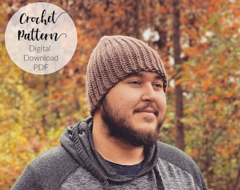 May include: A brown crochet beanie hat with a ribbed texture. The hat is being worn by a person with a beard, standing in front of a blurred background of fall foliage. The text "Crochet Pattern" is written in a white circle with a black outline. The text "Digital Download PDF" is written below the circle.