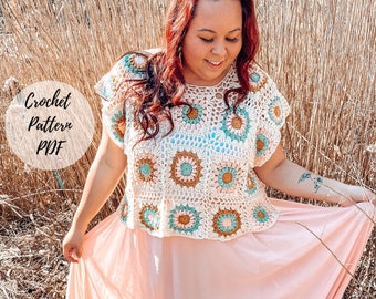 May include: A woman wearing a crocheted top with a floral pattern in shades of brown, pink, and teal. The top has a loose fit and short sleeves. The woman is standing in a field of tall grass.