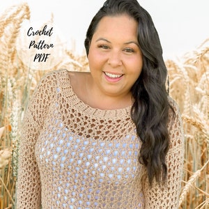 Puede incluir: Un top de crochet de corte holgado con un delicado patrón de calado. El top es de color beige claro y tiene mangas largas. La modelo lleva el top con unos vaqueros azules y está de pie en un campo de trigo.