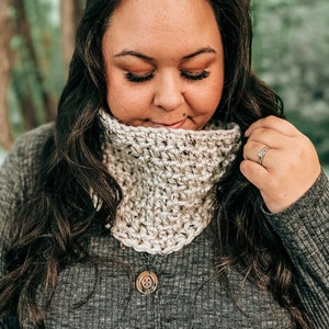 May include: A close-up of a woman wearing a beige crocheted cowl neck scarf. The scarf is made with a simple stitch pattern and has a relaxed, casual look.