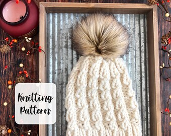 May include: A white knitted beanie with a brown faux fur pom pom. The beanie is sitting on a metal tray with a wooden frame. The text "knitting pattern" is written on a white square.