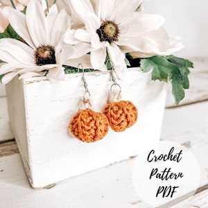 May include: Crochet pumpkin earrings in a brown and white colour scheme. The earrings are hanging from a white wooden box with a floral arrangement. The text "Crochet Pattern PDF" is visible in the image.