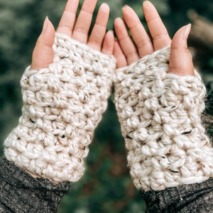 Könnte beinhalten: Ein Paar weiße, gehäkelte Fingerlose Handschuhe mit einem strukturierten Stichmuster. Die Handschuhe werden an den Händen einer Person getragen, wobei die Fingerspitzen frei bleiben.