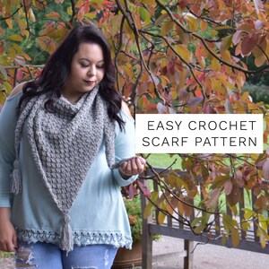 Puede incluir: Una mujer con una camisa azul claro y pantalones vaqueros sostiene una bufanda de ganchillo gris con borlas. La bufanda tiene forma triangular y un borde de encaje. El texto "EASY CROCHET SCARF PATTERN" está superpuesto a la imagen.