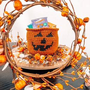 May include: A crocheted orange pumpkin-shaped basket with a black smiling face. The basket is filled with small bags of fruit snacks. The basket is sitting on a wooden plate surrounded by a fall-themed wreath with orange berries.