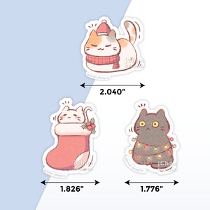May include: Three Christmas-themed cat stickers. The first sticker is a calico cat wearing a red Santa hat and scarf. The second sticker is a white cat with a red stocking and holly. The third sticker is a black cat with a string of Christmas lights. The stickers are approximately 2 inches wide.