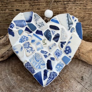 Può includere: Mosaico a forma di cuore realizzato con frammenti di ceramica blu e bianca. Il mosaico è montato su una base bianca e ha una corda di iuta per appenderlo. I pezzi di ceramica presentano vari motivi e sfumature di blu.