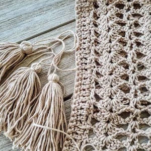 Peut inclure: Un gros plan sur un châle beige crocheté avec des franges. Le châle est réalisé avec un motif de point délicat et a une texture douce et naturelle.