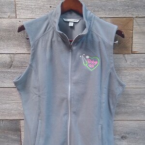 May include: Gray fleece vest with a zipper closure and a pink heart with a stethoscope and golf ball graphic that says "Robyn RN".