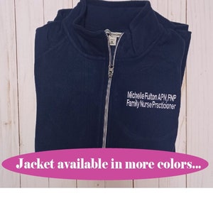 May include: A navy blue zippered jacket with a white embroidered name and title: "Michelle Fulton APN, FNP Family Nurse Practitioner". The text "Jacket available in more colors..." is displayed below the image.
