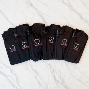 May include: Six black zip-up scrub jackets arranged in a row. Each jacket features an embroidered white tooth outline and a name with a professional title in pink thread, including Liz RDA, Crystal RDA, Christina RDA, Lizette, Jonal RDH, and Christina.
