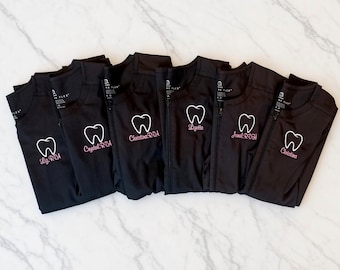 Op de afbeelding: Zes zwarte scrubjacks met rits op een rij. Elk jack is voorzien van een geborduurde witte tandcontour en een naam met professionele titel in roze draad, waaronder Liz RDA, Crystal RDA, Christina RDA, Lizette, Jonal RDH en Christina.