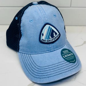 May include: A light blue baseball cap with a dark navy blue mesh back and a curved brim. The front features a triangular patch with a mountain graphic and the text "SKI CALIFORNIA". A circular sticker on the brim reads "DASHBOARD ADJUSTABLE LEGACY".