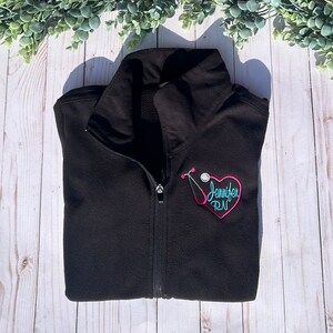 May include: Black fleece jacket with a pink heart and a stethoscope embroidered on the left chest. The heart has the text "Jennifer RN" embroidered inside.