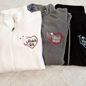 May include: Three fleece zip-up jackets with embroidered hearts and stethoscopes. The jackets are white, gray, and black. The hearts are red, blue, and pink. The text on the jackets reads "Michelle RN", "Heidi RN, BSN", and "Quanisho LPN".