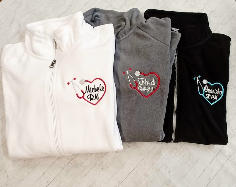 May include: Three fleece zip-up jackets with embroidered hearts and stethoscopes. The jackets are white, gray, and black. The hearts are red, blue, and pink. The text on the jackets reads "Michelle RN", "Heidi RN, BSN", and "Quanisho LPN".